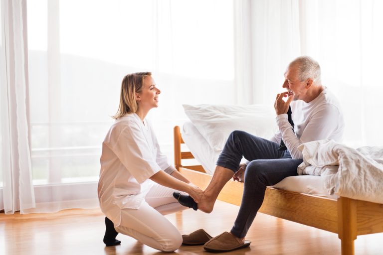 A female caregiver kneels down to help the old man on the bed to her right put on his black sock.