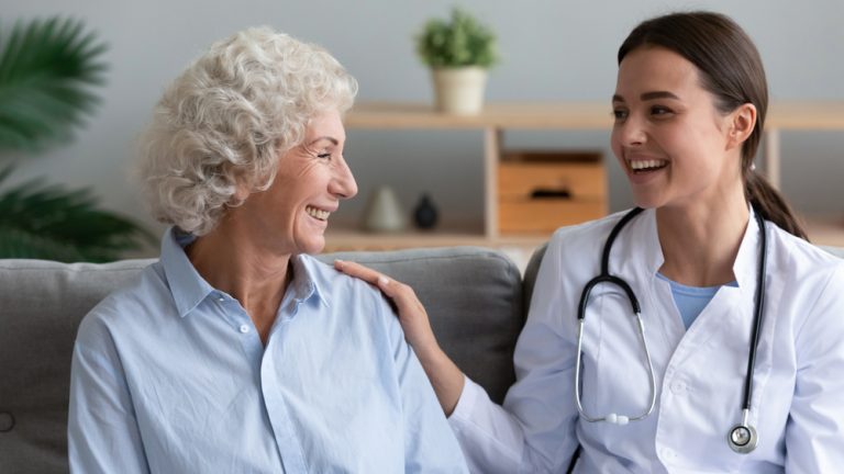 A female nurse and an old woman sit together on a couch, laughing and looking at each other. The nurse has one hand on the woman's shoulder.