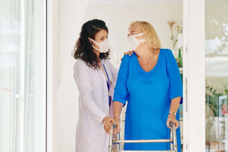 Two women look at each other and laugh. On the left is a nurse wearing a white lab jacket, and on the right is a woman in blue holding a walker. Both are wearing white facemasks.