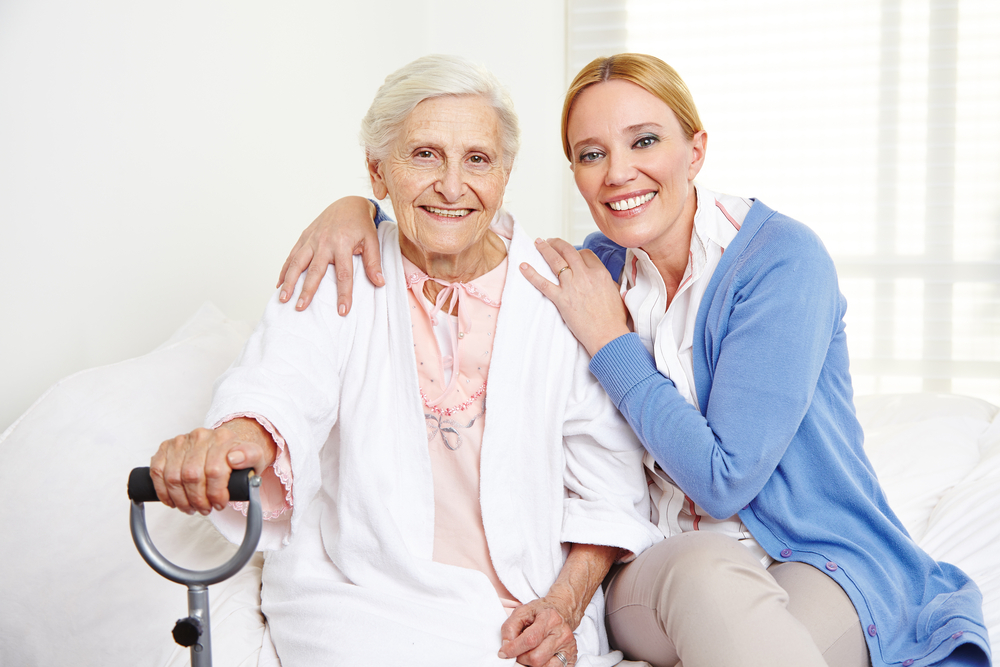 A female caregiver (right) poses with an elderly woman (left). The woman is wearing a blue jacket and the elderly woman is wearing a white one.