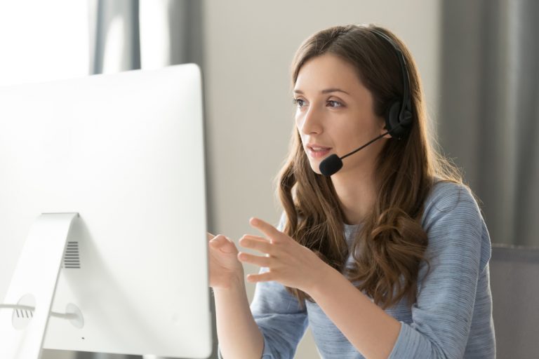 A woman with a phone headset talks to a customer while looking at her desktop computer in front of her.