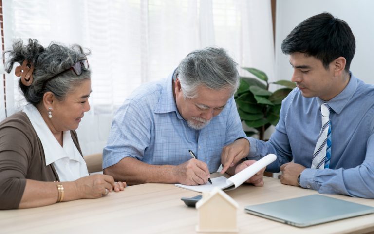 Two men and a woman sit at a table discussing a document placed in front of them. The man in the middle appears to be signing the document.