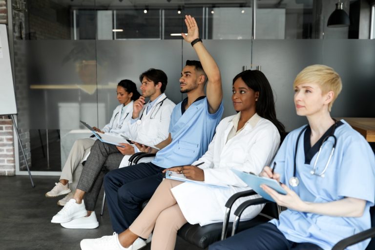 A group of five male and female nurses sits in a row, taking notes on a presentation off-screen. The man in the middle is raising his hand to ask a question.