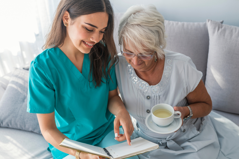 An elderly patient and a nurse sit on a couch. The patient has a mug of tea in her hand, and the nurse is reading her a story.