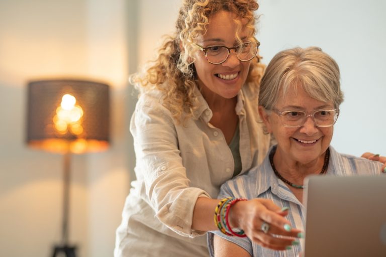 A caregiver and a patient look over a computer monitor. The patient is sat at a desk, while the caregiver is right behind her.