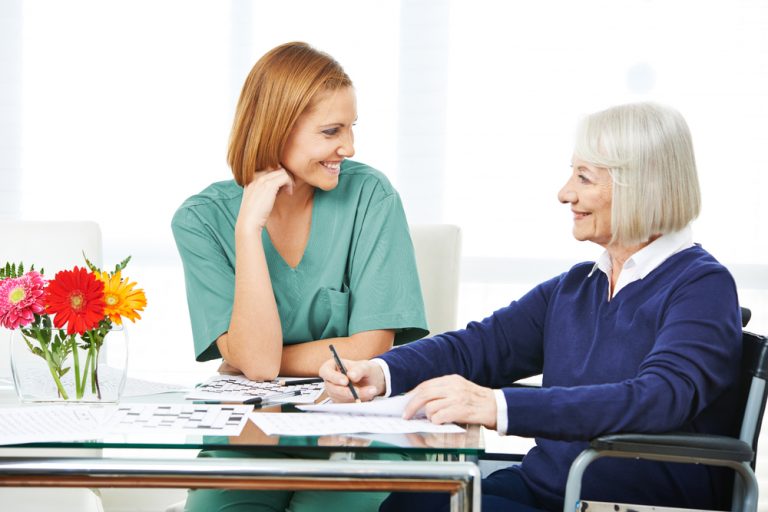 A caregiver sits with an older woman at a table. The woman is writing something on a piece of paper