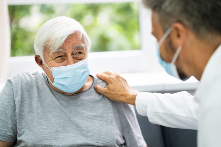 A doctor in a facemask off frame puts a hand on an elderly man's shoulder, who is looking at him. Both men are wearing facemasks